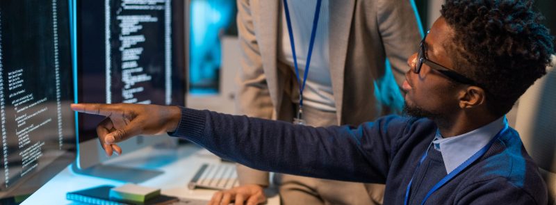 Two young intercultural colleagues discussing coded data on computer screen while African businessman pointing at it